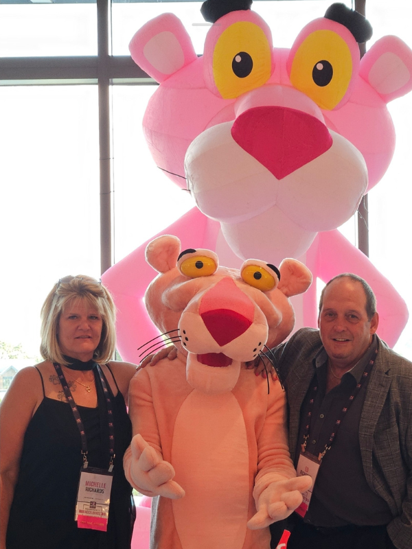 Michelle and Ed Richards smiling and posing with the CEEE, Owens Corning Pink Panther mascot.