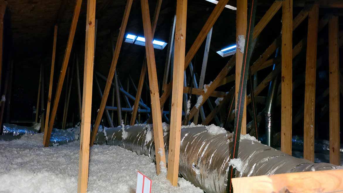 The interior of a FL home attic filled with thick, white blown-in insulation and large silver HVAC ducts running between wooden roof trusses.