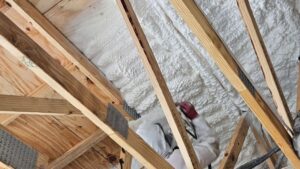A worker in protective gear applying white spray foam insulation between wooden roof rafters during new construction